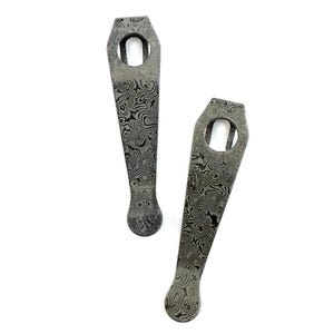 May include: Two metal pocket clips featuring a distinct dark gray and black swirling Damascus steel pattern. Each clip has a tapered body, a rounded tip, and a circular mounting hole at the wider top end, showcasing a complex layered metal texture.