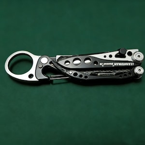 May include: A black and silver multi-tool with a carabiner clip and the text "LEATHERMAN SKELETOOL KB" on a dark green background. The tool has a variety of features, including a knife blade and a bottle opener.