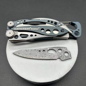 Puede incluir: Una multiherramienta Leatherman Skeletool plateada y azul, y una hoja separada con diseño. La multiherramienta está cerrada y la hoja descansa sobre una superficie circular blanca. El logotipo de Leatherman es visible.