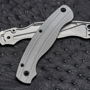 May include: Two grey metal knife handle scales. One scale features a textured, cross-hatched pattern with three circular holes. The second scale is a smooth, flat piece of grey metal with cutouts and circular holes for assembly and mounting.
