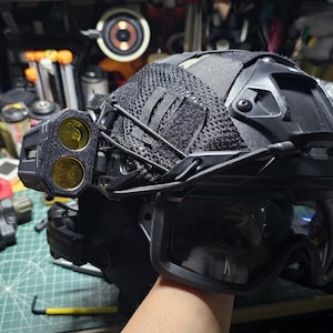 Könnte beinhalten: Schwarzer taktischer Helm mit angebrachter Schutzbrille, einem Netzbezug und einem Doppellichtsystem mit gelben Linsen. Der Helm wird vor einer unordentlichen Werkbank gehalten, was auf militärische oder taktische Ausrüstung hindeutet.