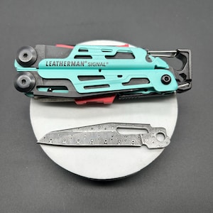 Könnte beinhalten: Ein türkis-schwarzes Multi-Tool mit dem Text "LEATHERMAN SIGNAL". Eine separate Messer-Klinge mit Muster ist ebenfalls sichtbar. Das Multi-Tool hat einen roten Akzent und verschiedene Werkzeuge. Die Artikel befinden sich auf einer weißen Oberfläche.