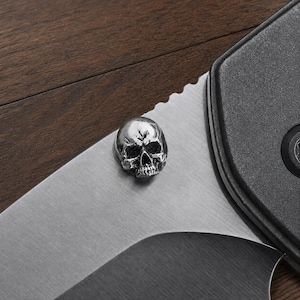 May include: A small, three-dimensional silver-coloured metal skull bead or stud attached to the flat, brushed steel blade of a folding knife. The skull features detailed eye sockets, a nasal cavity, and teeth, with a polished metallic finish.