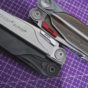 Könnte beinhalten: Zwei Multitools, eines in Schwarz und eines in Silber, werden auf einer lila Gitteroberfläche gezeigt. Das silberne Werkzeug hat einen roten Akzent. Der Text "LEATHERMAN SURGE" ist auf beiden Werkzeugen sichtbar.