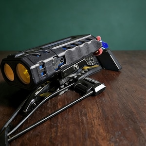 Könnte beinhalten: Ein schwarzes und silbernes Gerät mit zwei gelben Linsen, möglicherweise eine Kamera oder ein wissenschaftliches Instrument, liegt auf einer Holzoberfläche. Das Gerät hat ein komplexes Design mit mehreren Komponenten und einem Griff. Der Hintergrund ist einfarbig grün.