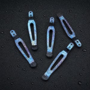 May include: Four elongated metal pocket clips with a textured, iridescent blue and purple finish. Each clip features a narrow, open-center design, a rounded tip, and two small circular holes at the top for attachment, arranged in a scattered pattern.