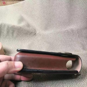 Puede incluir: Una funda de cuero marr&oacute;n con ribete negro y una abertura circular. La funda est&aacute; sujeta por una mano y parece ser una funda protectora para un dispositivo. La funda tiene un dise&ntilde;o curvo y un cierre met&aacute;lico.