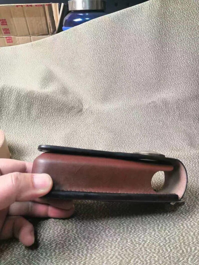 Puede incluir: Una funda de cuero marr&oacute;n con ribete negro y una abertura circular. La funda est&aacute; sujeta por una mano y parece ser una funda protectora para un dispositivo. La funda tiene un dise&ntilde;o curvo y un cierre met&aacute;lico.