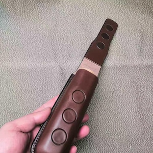Puede incluir: Una funda de cuchillo de cuero marr&oacute;n con tres hendiduras circulares en la parte delantera y tres hendiduras circulares m&aacute;s peque&ntilde;as en la parte superior. La funda se sostiene en una mano sobre un fondo texturizado.