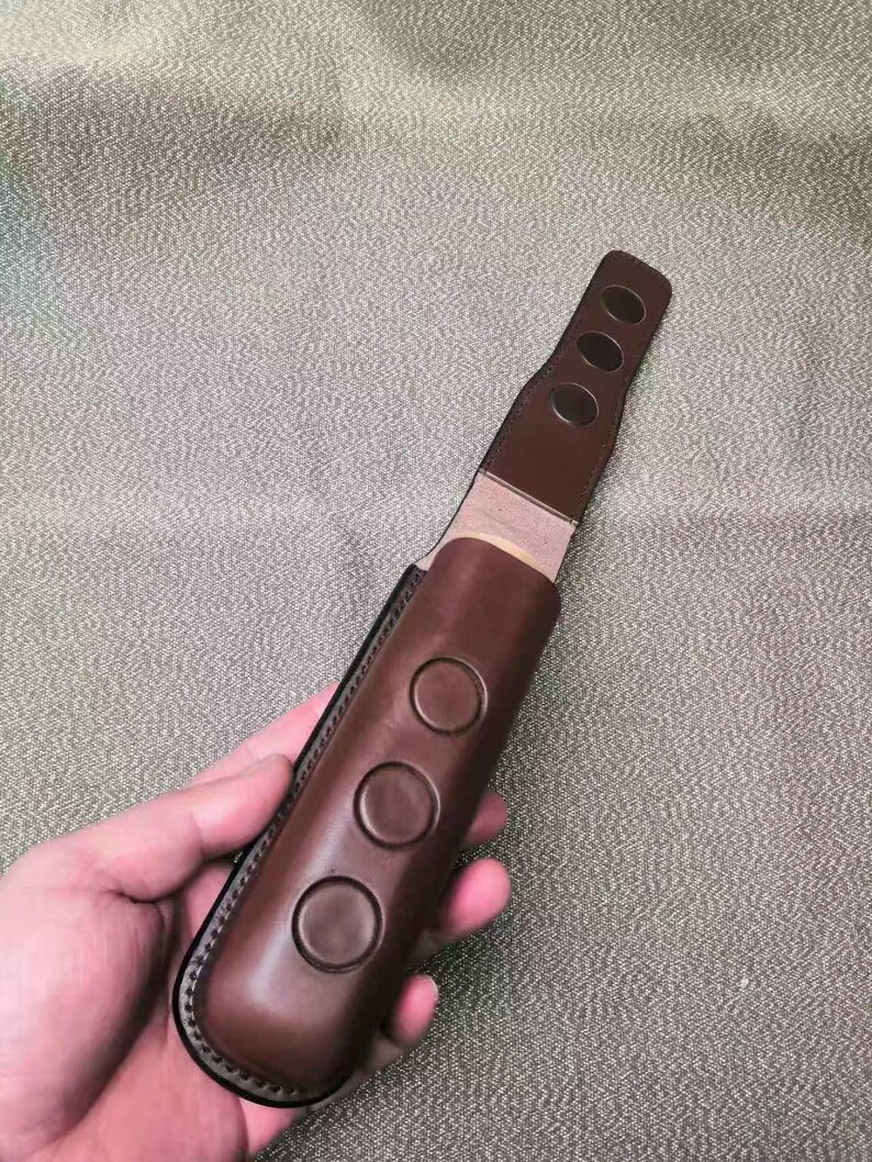 Puede incluir: Una funda de cuchillo de cuero marr&oacute;n con tres hendiduras circulares en la parte delantera y tres hendiduras circulares m&aacute;s peque&ntilde;as en la parte superior. La funda se sostiene en una mano sobre un fondo texturizado.
