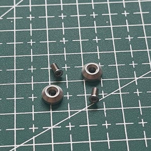 以下が含まれることがあります： 2つの銀色の丸みを帯びた金属ワッシャーと2つの小さなネジのクローズアップ。ワッシャーには中央の穴と面取りされたエッジがあります。ネジは六角穴付きです。緑色のカッティングマットの上にあります。