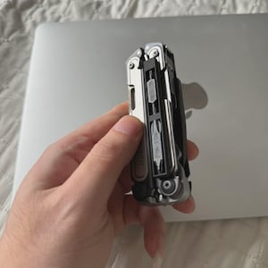 May include: A multi-tool with a silver and black finish, featuring various tools like a knife, pliers, and screwdrivers. The tool is partially open, revealing its components. The word "LEATHERMAN" is visible on the side.