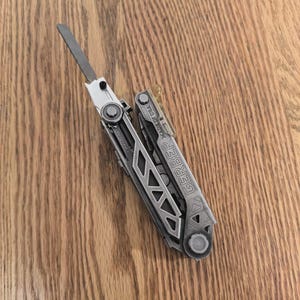 Pode incluir: Uma caneca de cerâmica branca com uma alça preta e o texto "But First, Coffee" em negrito preto. A caneca está sobre uma superfície branca. A caneca tem aproximadamente 10 cm de altura.