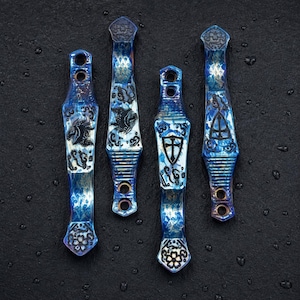 Puede incluir: Cuatro clips de bolsillo metálicos alargados con un acabado iridiscente azul y morado. Cada clip presenta intrincados diseños grabados con láser en negro, incluyendo patrones florales, un escudo con una cruz y formas abstractas, con dos orificios.