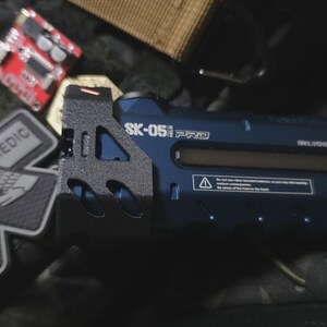 Könnte beinhalten: Ein dunkelblaues SK-051 PRO-Gerät mit einem schwarzen, strukturierten Griff, einem transparenten Display und weißem Text. Das Bild zeigt auch eine rote Leiterplatte, einen grau-weißen Medic-Patch mit einem Totenkopf und andere taktische Ausrüstung.