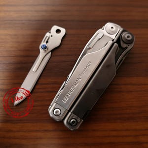Könnte beinhalten: Ein silbernes Leatherman Surge Multitool in geschlossener Position neben einer separaten silbernen Metallklinge mit einer kleinen blauen Pivot-Schraube. Der Markenname und das Modell sind auf der Seite des Griffs des Multitools eingraviert.