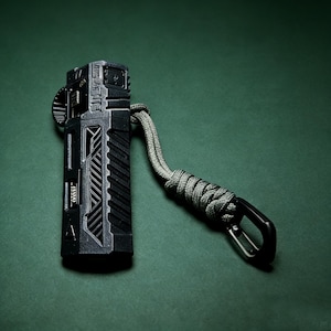 以下が含まれることがあります： 黒色の工業用ライター。テクスチャード加工のデザインで、グレーのストラップが黒いカラビナクリップに付いています。ライターは複雑な幾何学模様で、濃い緑色の背景に配置されています。