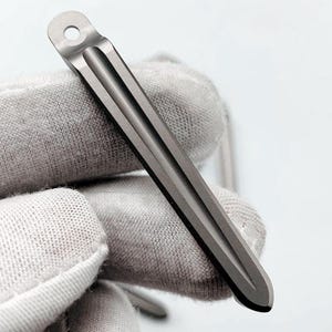 May include: A close-up of a small, gray metal tool. The tool has a flat, rectangular body with a rounded tip and a hole at the top. It features three parallel grooves running down its length. The tool is held by a gloved hand.