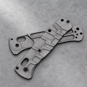 May include: Two dark gray metal knife handle scales featuring a textured, weathered finish. Each scale has a series of rectangular cutouts, circular screw holes, and a larger oval opening, designed for assembly onto a folding knife frame.