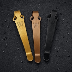 Puede incluir: Tres clips de bolsillo de metal en acabados dorado, bronce y negro. Cada clip presenta un diseño cónico con dos orificios de montaje circulares en la parte superior y un pequeño icono de mariposa grabado en la superficie del cuerpo metálico.
