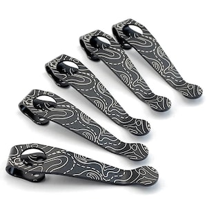 May include: Five black metal pocket clips with a white topographical pattern. Each clip has a curved design with a hole at one end and a rounded tip at the other. The clips are arranged on a white background.