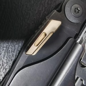 Könnte beinhalten: Nahaufnahme eines Multitools mit silberner und goldener Oberfläche. Das Werkzeug verfügt über eine Klinge, einen Schraubendreher und andere Werkzeuge. Das goldfarbene Element hat eine rechteckige Form mit einem Ausschnitt. Das Werkzeug ist für verschiedene Aufgaben konzipiert.