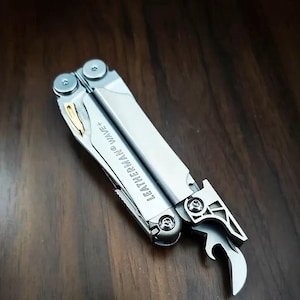 以下が含まれることがあります： シルバーカラーのLeatherman Wave+マルチツール。メタリックな質感でゴールドのアクセントが施されています。ツールは部分的に展開されており、ボトルオープナーと、ハンドルに目に見えるネジで固定された金属製のコンポーネントが確認できます。