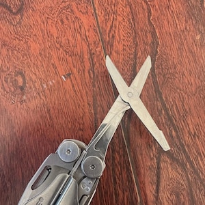 May include: A silver multi-tool with scissors extended. The tool is partially open, showing the scissors. The handle is engraved with the word "WAVE". The background is a wooden surface.