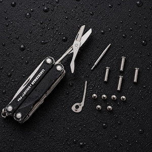 Może przedstawiać: Czarne narzędzie wielofunkcyjne Leatherman Squirt PS4 z rozłożonymi nożyczkami, w towarzystwie kilku małych metalowych szpilek, nitów i cienkiego metalowego elementu. Narzędzie posiada srebrne akcenty i jest pokazane wraz z różnymi zdemontowanymi częściami.