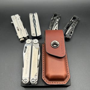 May include: A selection of multi-tools, including a Leatherman Wave, presented with a brown leather sheath. The tools are silver and black, featuring folding components. The sheath has a snap closure and the Leatherman logo.
