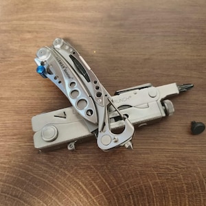 May include: A silver multi-tool with various functions, including pliers and a screwdriver. The tool is partially open, displaying its features. A small black component is positioned nearby. The tool rests on a wooden surface.