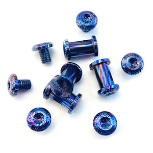 May include: A selection of blue and purple metal body jewellery, including screw-on plugs and flat-topped screws. The jewellery has a swirling, patterned design. The pieces are scattered on a white surface.