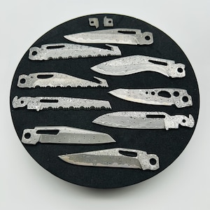May include: An assortment of unfinished, patterned metal knife blades and small parts are arranged on a black circular surface. The blades vary in shape, including straight, curved, and serrated edges. The metal has a unique, swirling pattern.