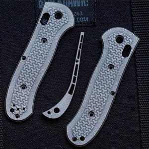 May include: Two grey textured knife handle scales with a repeating geometric pattern and a matching curved backspacer. Each scale features multiple circular screw holes and a larger pivot hole, designed for assembly on a folding pocket knife frame.