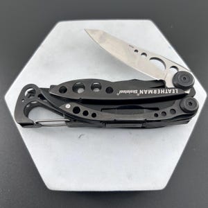 Puede incluir: Una multiherramienta Leatherman Skeletool negra con una hoja abierta. La herramienta cuenta con un mosquetón y el nombre de la marca es visible. La hoja es plateada y la herramienta está sobre una superficie blanca.