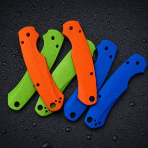 Op de afbeelding: Een set van vijf handgrepen voor messen met getextureerde oppervlakken en voorgeboorde gaten. De set bevat twee feloranje, één limoengroene en twee felblauwe handgrepen, die in een overlappend patroon zijn gerangschikt.