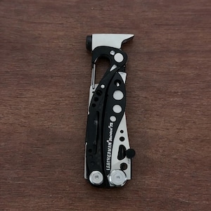 May include: A black and silver Leatherman multi-tool, featuring a variety of tools. The multi-tool is shown open, displaying its components. The Leatherman logo is visible. The tool is designed for various tasks and is made of metal.