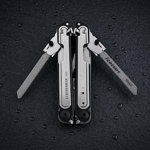 Könnte beinhalten: Ein silber-schwarzes Leatherman Arc Multitool mit zwei ausgeklappten Sägeblättern. Ein Sägeblatt ist mit dem Text T-SHANK und Metal beschriftet. Das Werkzeug hat eine gebürstete Metalloberfläche an den Griffen und schwarze Akzente an den Gelenken.