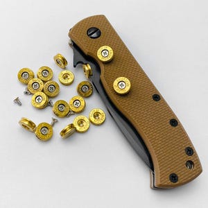 May include: A brown folding knife with a textured handle and black details. Several gold-coloured bullet casings with the text "EMERSON 45 AUTO" are scattered around the knife, along with small screws.