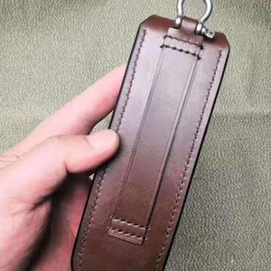 Puede incluir: Una funda de cuero marr&oacute;n con un anillo de metal en la parte superior. La funda tiene una trabilla en la parte trasera y est&aacute; cosida con un hilo en contraste. El cuero es de un color marr&oacute;n oscuro intenso.