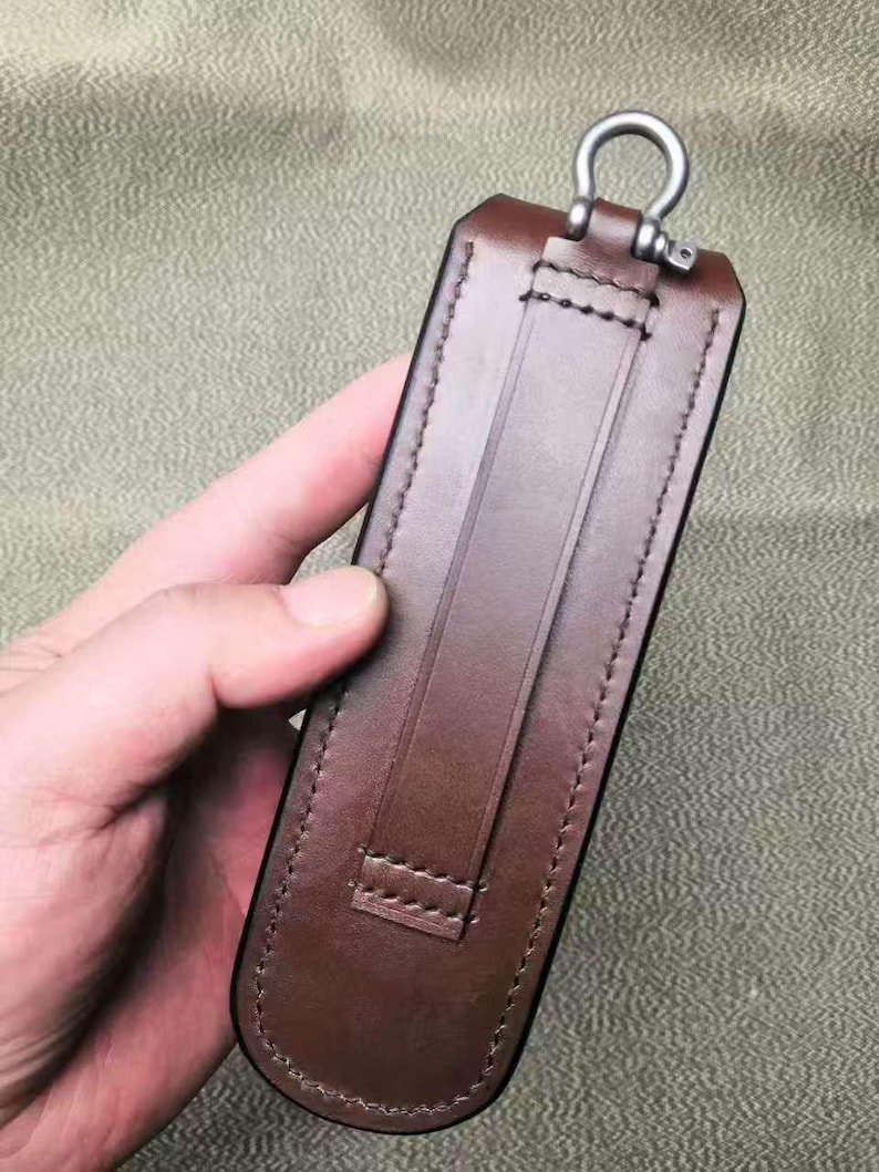 Puede incluir: Una funda de cuero marr&oacute;n con un anillo de metal en la parte superior. La funda tiene una trabilla en la parte trasera y est&aacute; cosida con un hilo en contraste. El cuero es de un color marr&oacute;n oscuro intenso.