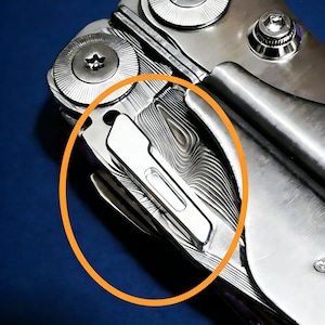Könnte beinhalten: Ein silberfarbenes Multifunktionswerkzeug mit einem markanten Taschenclip an einem Griff mit einem ausgeprägten wellenförmigen, geschichteten Metallmuster. Das Werkzeug verfügt über kreisförmige Pivot-Schrauben mit sternförmigen Zentren.