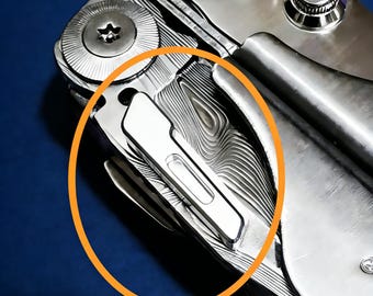 May include: Close-up of a multi-tool with a silver finish and a patterned design. The tool features a blade and other implements, with visible screws and a clip. The background is a solid blue.