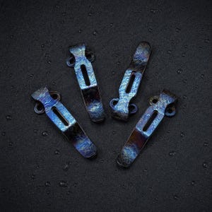 May include: Four metal pocket clips with a heat-anodized finish displaying a mottled pattern of deep blue, purple, and dark bronze tones. Each clip features a central elongated slot and two circular mounting holes at the top attachment point.