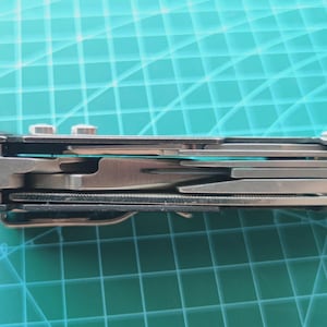Op de afbeelding: Een zilverkleurig multitool met diverse gereedschappen, waaronder een mes en een tang. Het gereedschap is gedeeltelijk open, waardoor de verschillende onderdelen zichtbaar zijn. De achtergrond is een turquoise snijmat.
