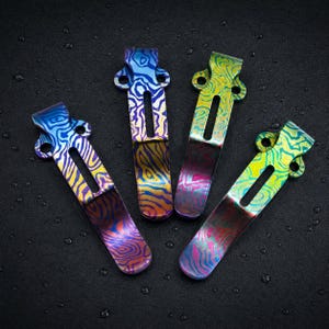 Puede incluir: Cuatro clips de bolsillo de metal con un acabado anodizado multicolor vibrante y un patrón topográfico ondulado. Cada clip tiene una sección superior curva con dos orificios de montaje y una ranura vertical estrecha en el centro.