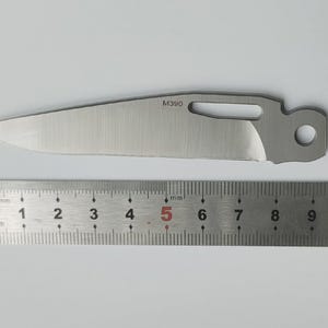 May include: A stainless steel knife blade with a partially sharpened edge, a hole, and the text "M390". A metal ruler with millimeter and centimeter markings is below the blade.