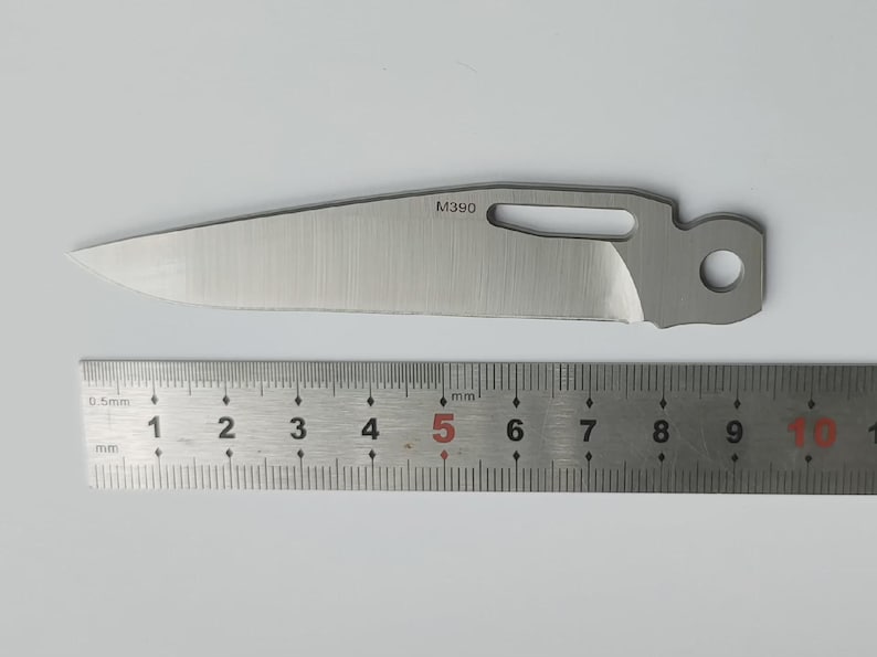 May include: A stainless steel knife blade with a partially sharpened edge, a hole, and the text "M390". A metal ruler with millimeter and centimeter markings is below the blade.