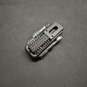 May include: A compact black multi-tool featuring a central row of integrated screwdriver bits, a side-mounted silver metal driver shaft with a grey collar, and a rectangular cutout handle frame with visible screw fasteners on the top surface.