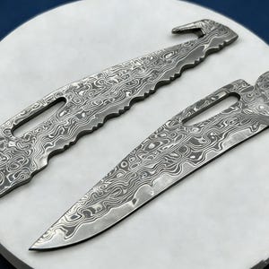 May include: Two metal tools with a distinctive patterned design. One has a serrated edge and a hook, the other a pointed blade. Both have a hole for hanging. The tools are displayed on a white circular surface.